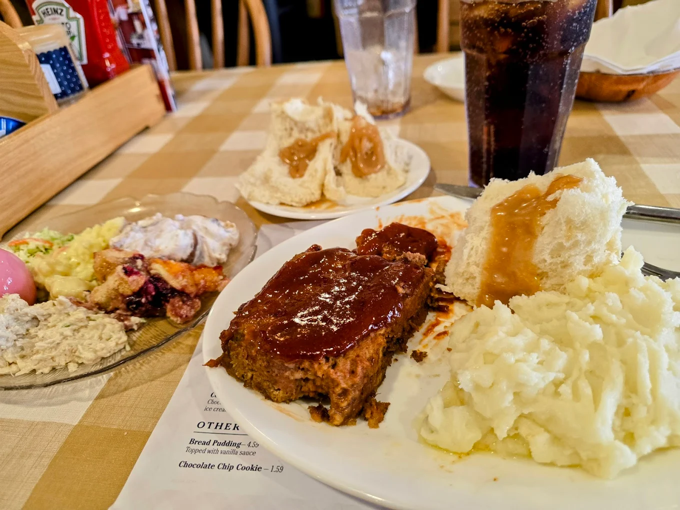 A plate with meatloaf topped with sauce, mashed potatoes with gravy, and a dinner roll sits on a checkered table. Also visible are a drink, dessert plate, and another plate with biscuits and gravy.