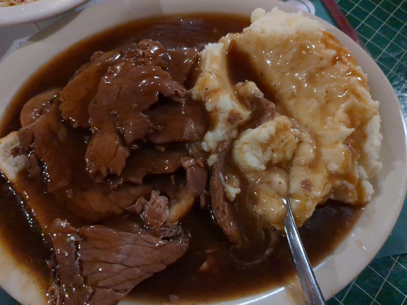A plate of sliced roast beef covered in brown gravy, served with a large portion of mashed potatoes and gravy, with a fork resting on the side.