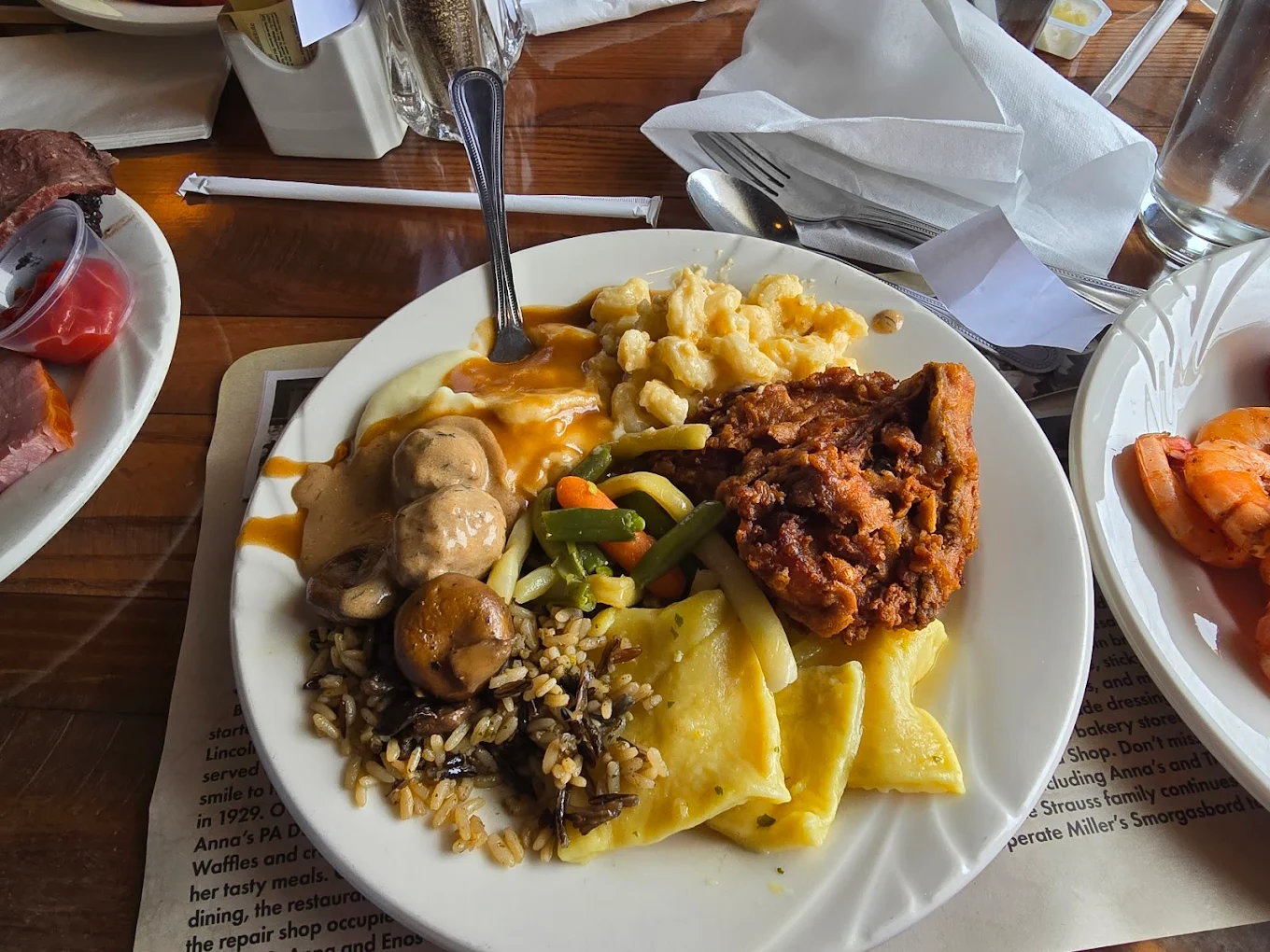 A white plate with macaroni and cheese, a breaded fried chicken piece with sauce, mixed vegetables, wild rice with mushrooms, two triangular pasta pieces, and brown gravy, on a wooden table with cutlery and a glass nearby.