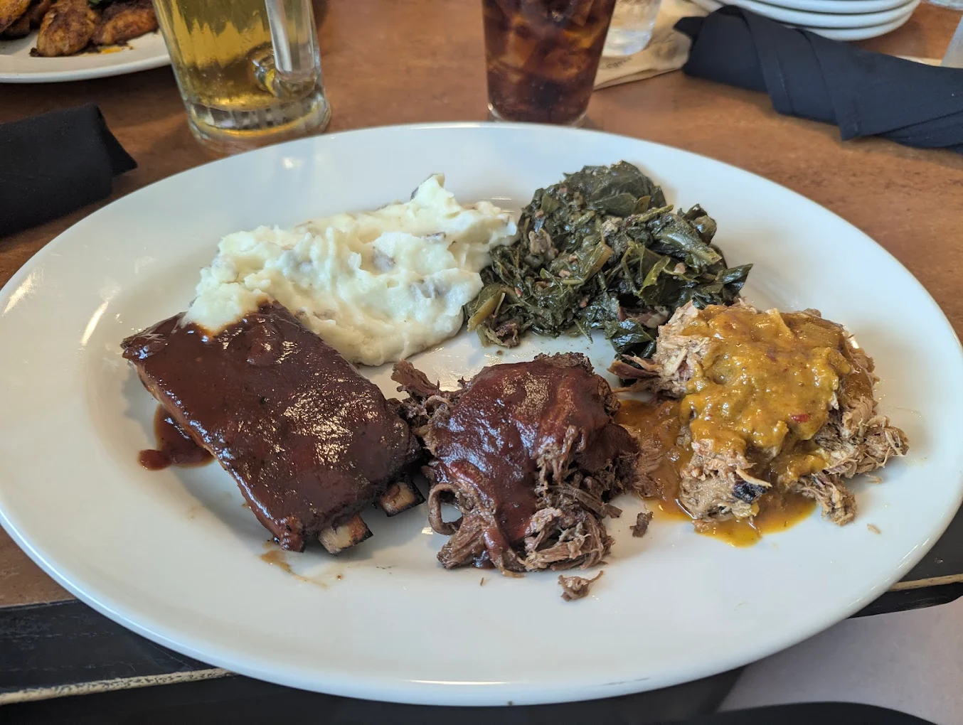 A white plate with mashed potatoes, cooked leafy greens, barbecue ribs, pulled beef with sauce, and pulled pork with a mustard-based sauce. Drinks and napkins are visible in the background.