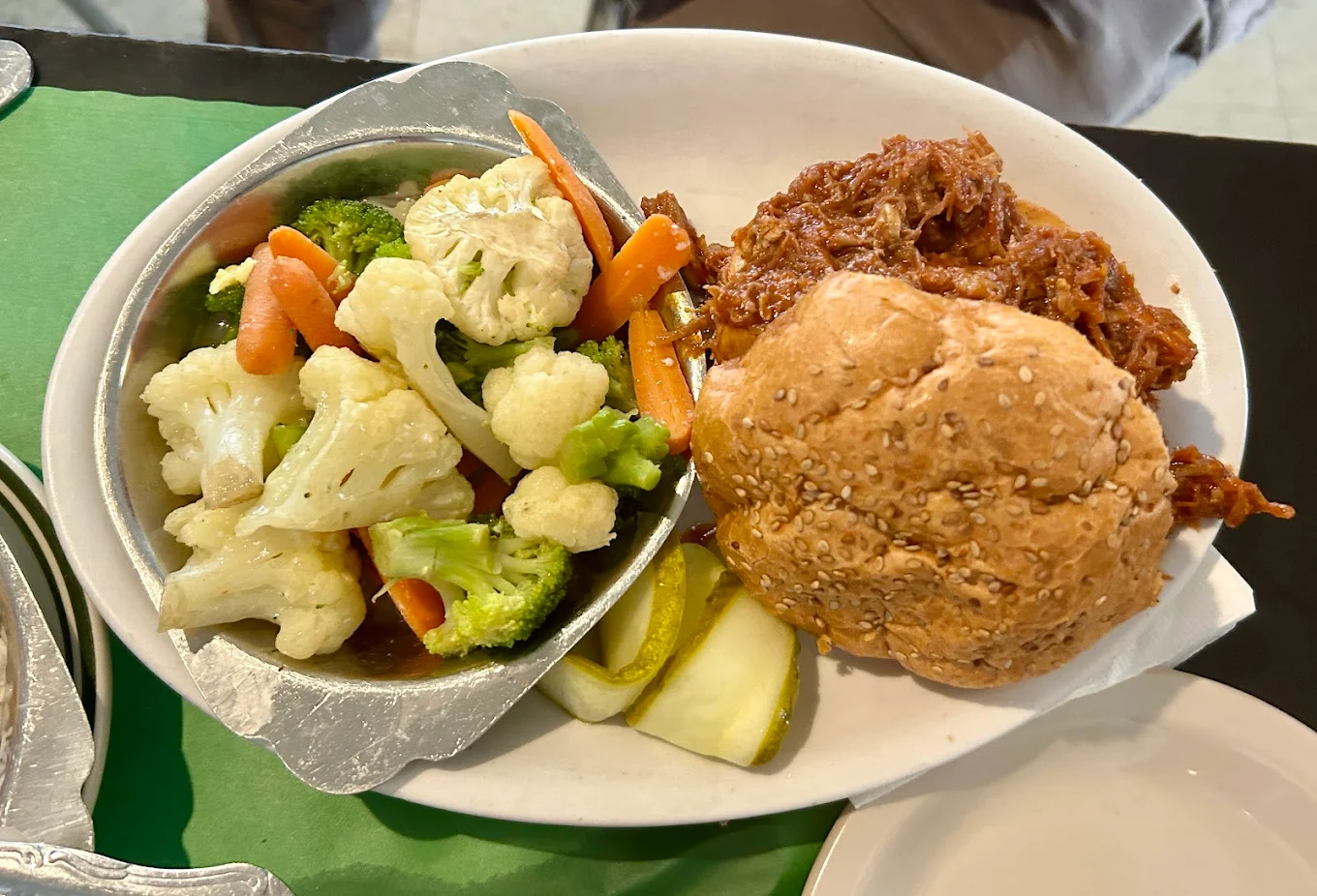 A plate with a pulled pork sandwich on a sesame bun, a side of mixed steamed vegetables (including broccoli, cauliflower, and carrots), and pickle slices.