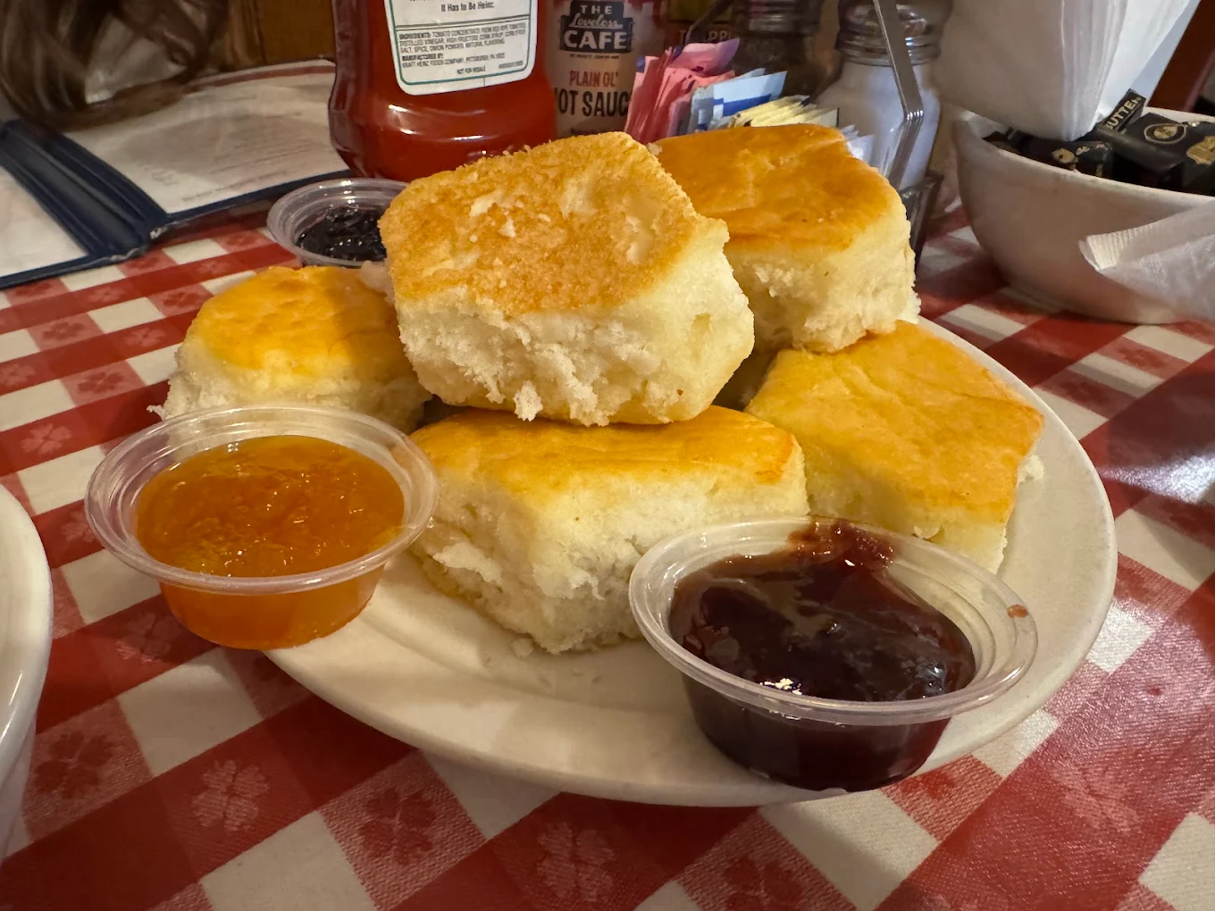 A plate of six golden, fluffy biscuits is served with two containers of orange and red fruit preserves on a red and white checkered tablecloth. Condiments and menus are visible in the background.