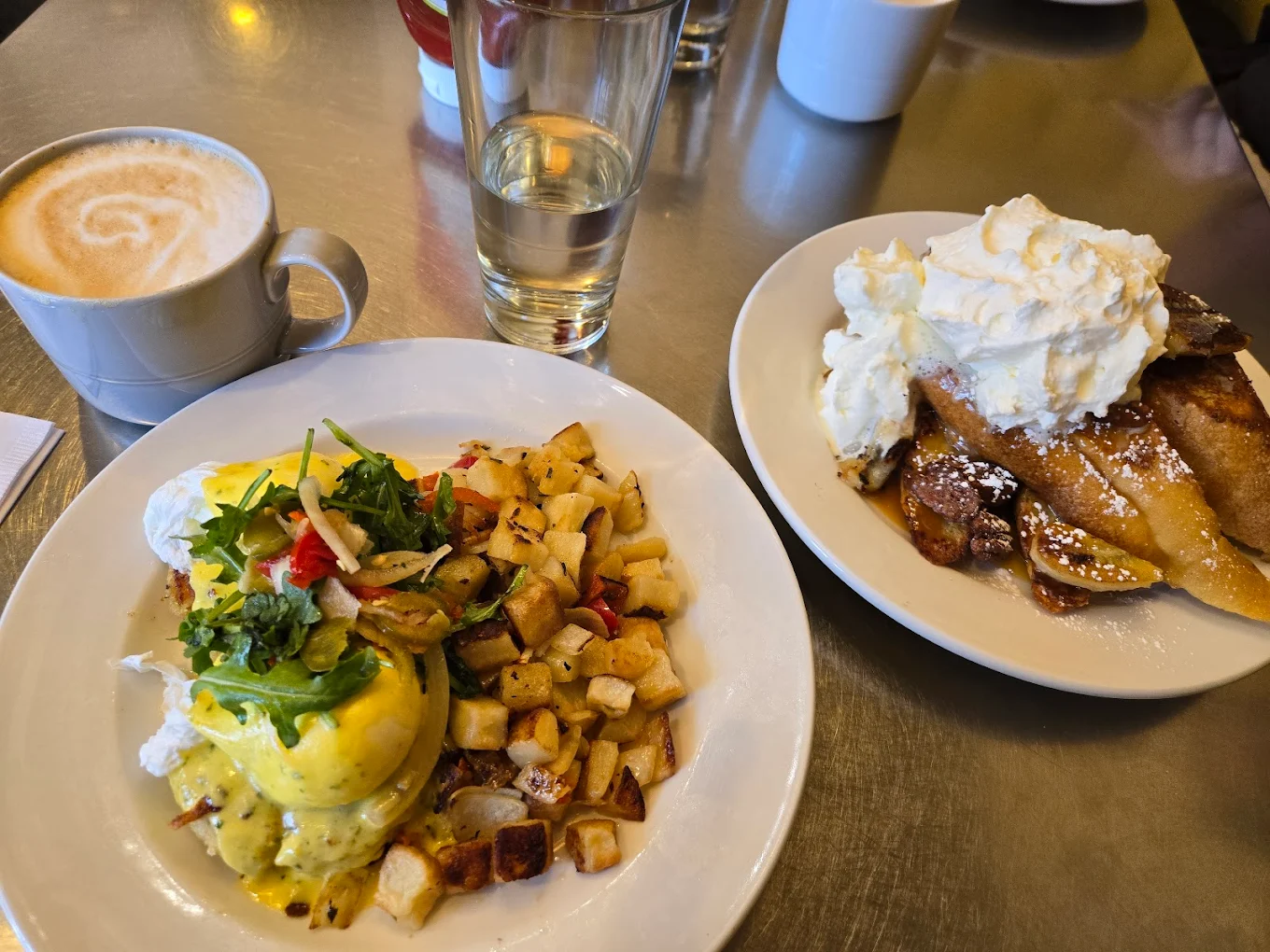 A breakfast spread with a plate of eggs Benedict, home fries, a cup of coffee, a glass of water, and a plate of French toast topped with whipped cream and bananas on a metal table.
