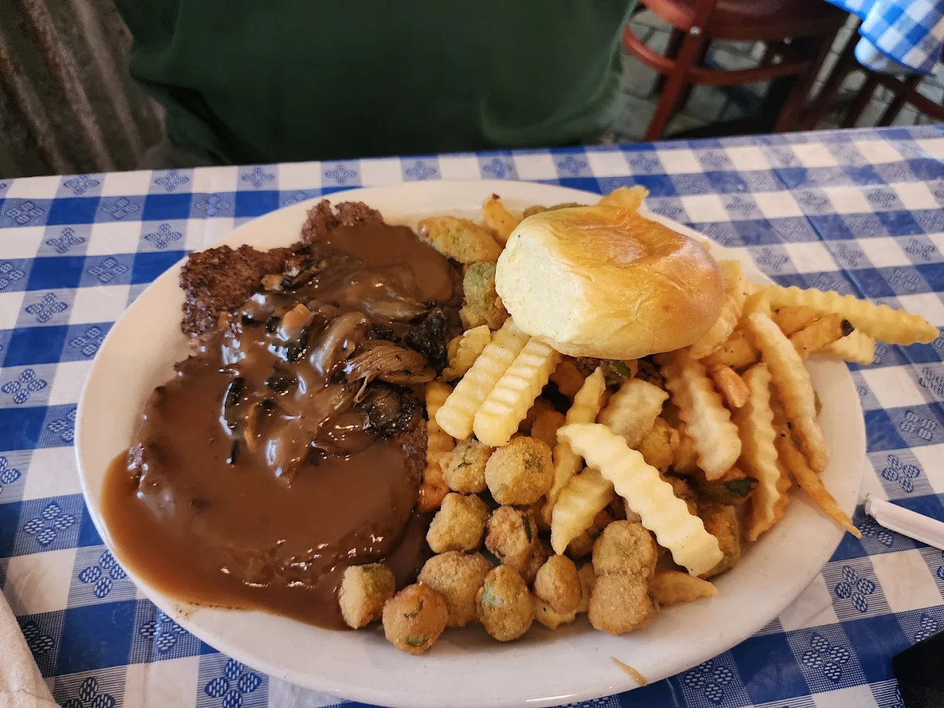 A plate of food on a blue and white checkered tablecloth with crinkle-cut fries, fried okra, a biscuit, and hamburger steak topped with brown gravy and onions.
