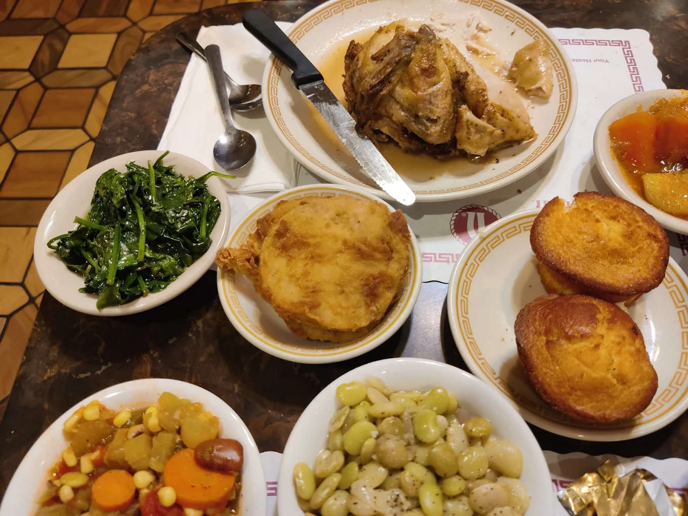 A table with plates of fried chicken, cornbread muffins, sautéed greens, fried green tomato, lima beans, a vegetable medley, apple sauce, and utensils on a patterned brown surface.