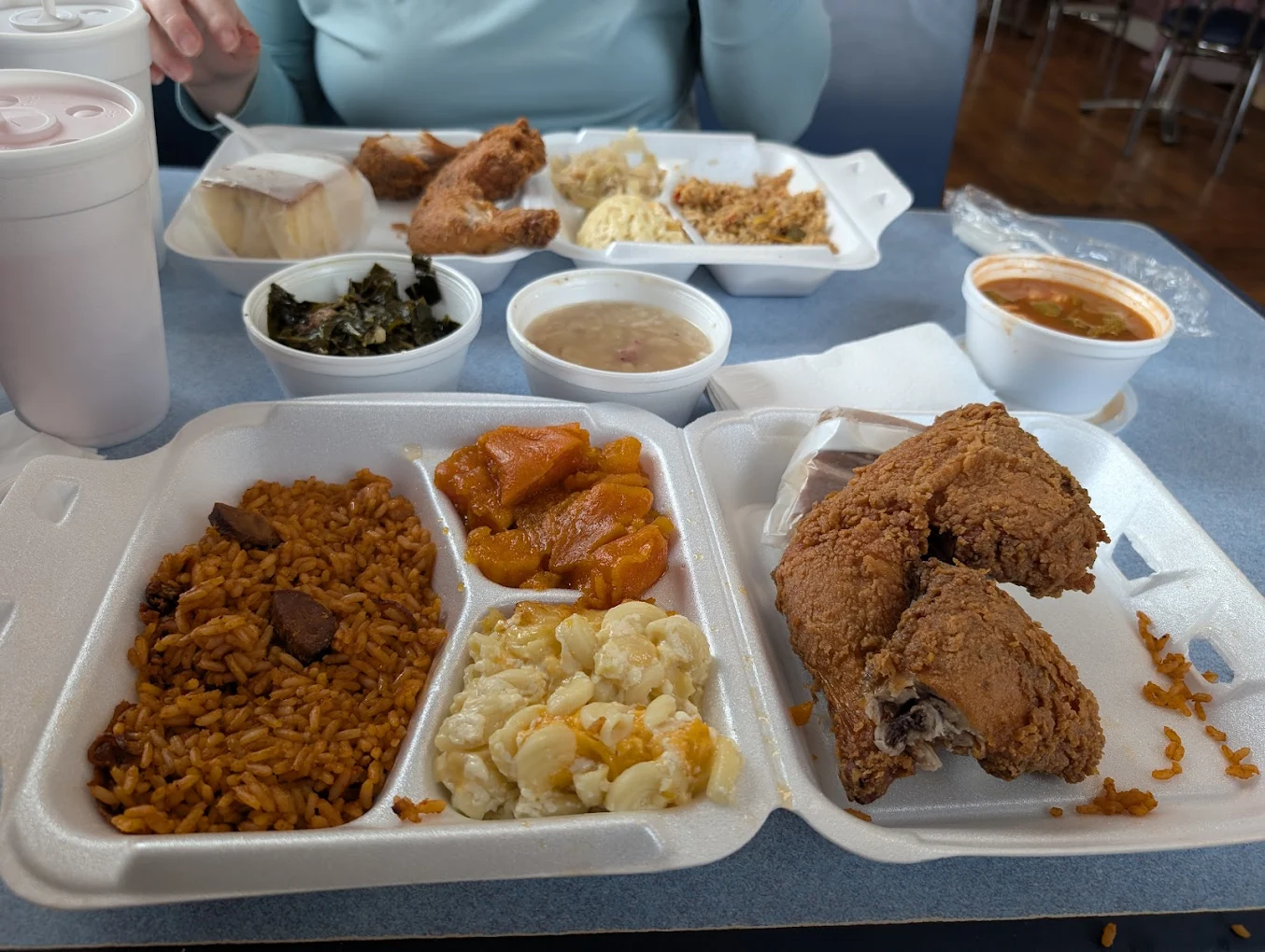 Two foam trays filled with fried chicken, macaroni and cheese, seasoned rice, and candied yams. Sides of collard greens, cornbread, stuffing, and drinks are also visible on a table with another person's meal.