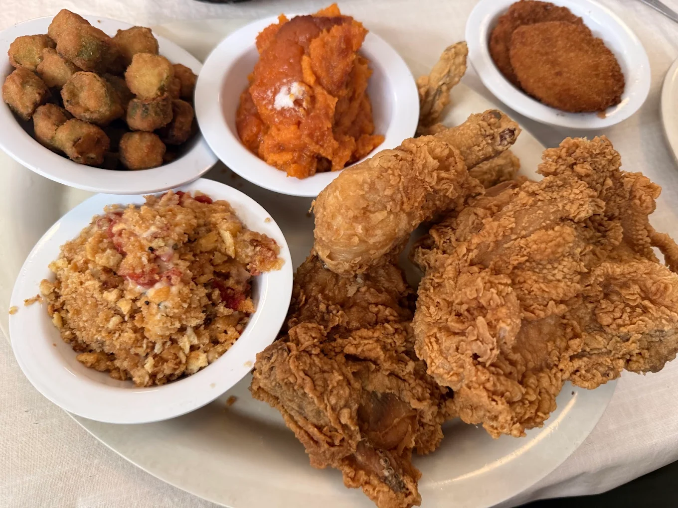 A plate of crispy fried chicken with side dishes in white bowls, including fried okra, sweet potatoes, cornbread dressing, and a breaded patty, all arranged on a white tablecloth.