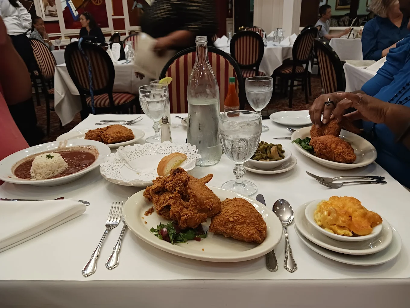 A restaurant table set for two with plates of fried chicken, macaroni and cheese, green beans, a bowl of rice with beans, bread, glasses of water, and a bottle of water, with people dining in the background.