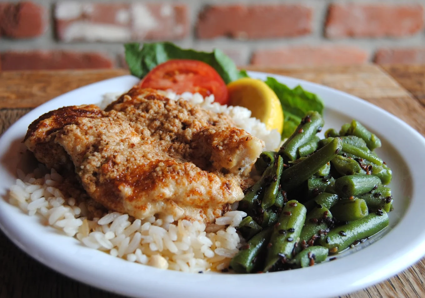 A plate with baked chicken over white rice, served with seasoned green beans and garnished with tomato, lettuce, and a lemon slice. A brick wall is visible in the background.