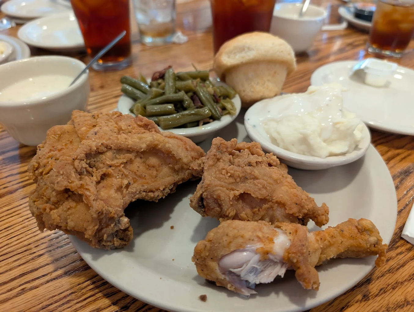 A plate with fried chicken, mashed potatoes with gravy, green beans, and a bread roll on a wooden table. Glasses of iced tea and bowls of sauce are in the background.