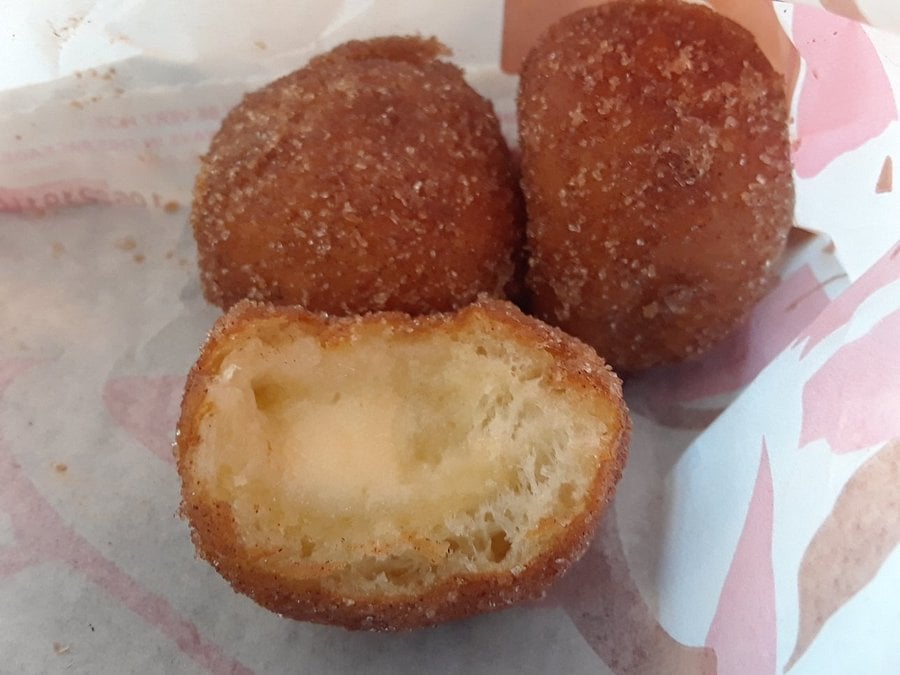 Three round, golden-brown doughnuts coated in cinnamon sugar, with one cut open to reveal a soft, airy inside. They are placed on white paper with a pink pattern.