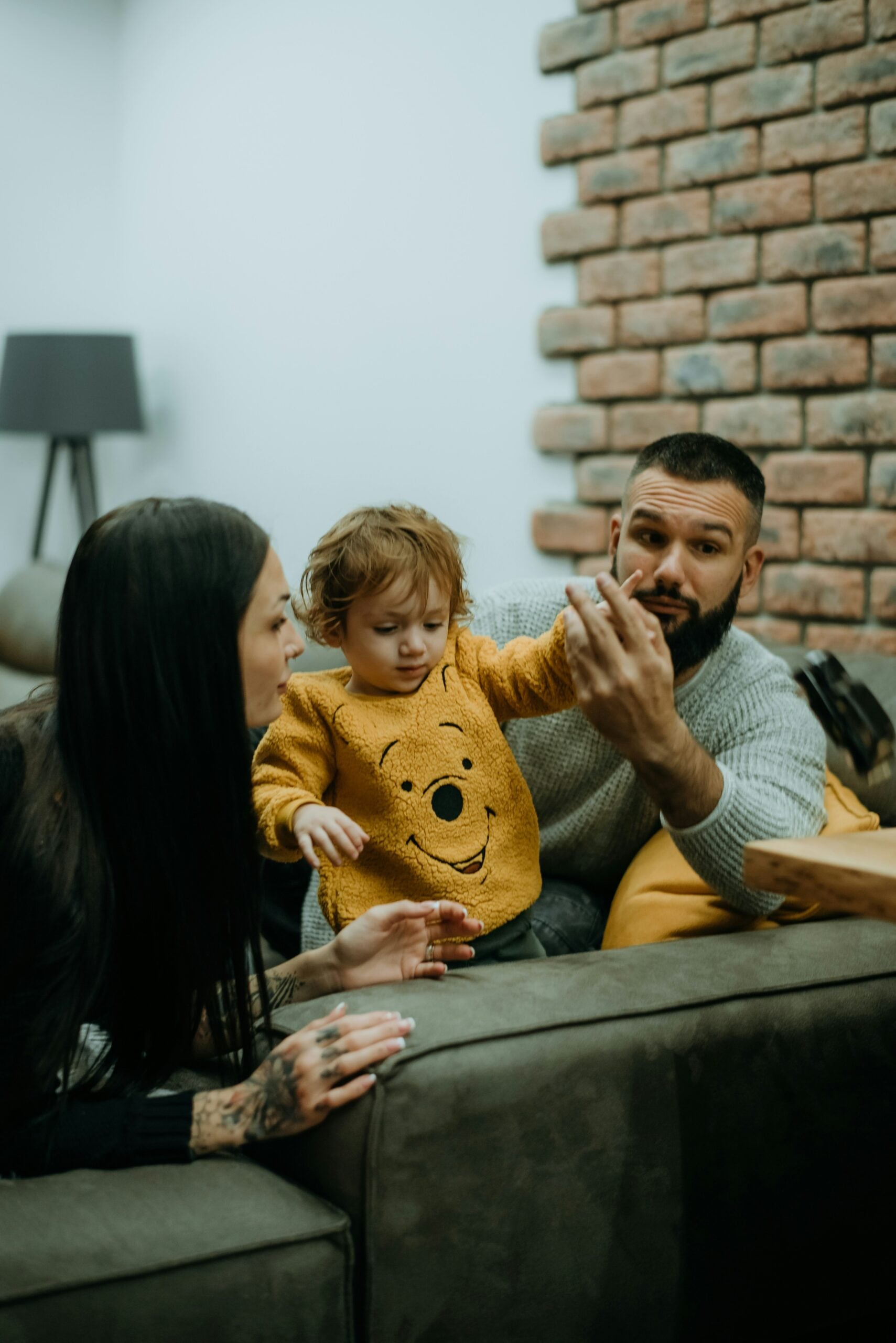 A man, woman, and toddler sit on a couch. The man holds a phone, showing it to the child, who wears a yellow sweater with a bear face. The woman leans in, watching the child. There’s a brick wall and lamp in the background.