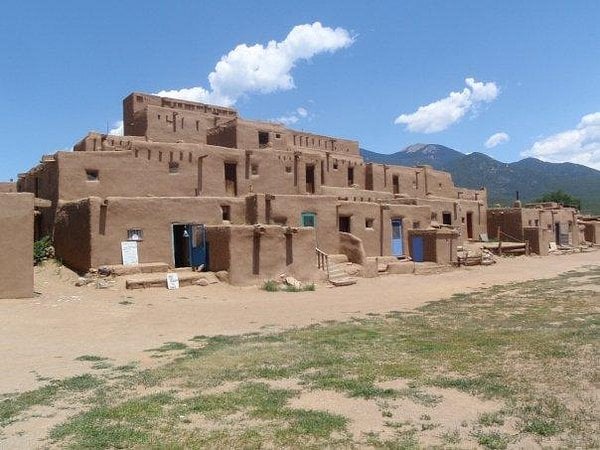 A large, multi-story adobe pueblo with tan mud walls and wooden beams, set against a blue sky and mountains—a striking example of Unique American Towns, featuring colorful doors and windows above sandy ground with grassy patches.