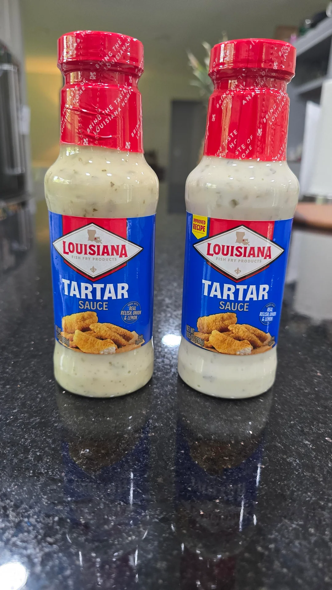 Two bottles of Louisiana Tartar Sauce with red caps and blue labels are placed side by side on a shiny black countertop. The labels feature a photo of fried food pieces.