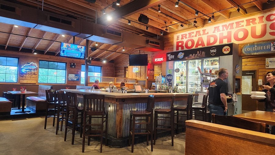 A rustic bar and grill interior with wooden walls and ceiling, a central bar surrounded by empty chairs, a few customers seated, TVs on the walls, and a large sign reading "REAL AMERICAN ROADHOUSE.