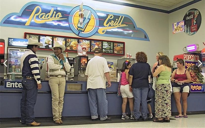 A group of people, including two men in cowboy hats, stand in line at a Radio Grill fast food counter inside a store, with a colorful sign and menu boards above the counter.