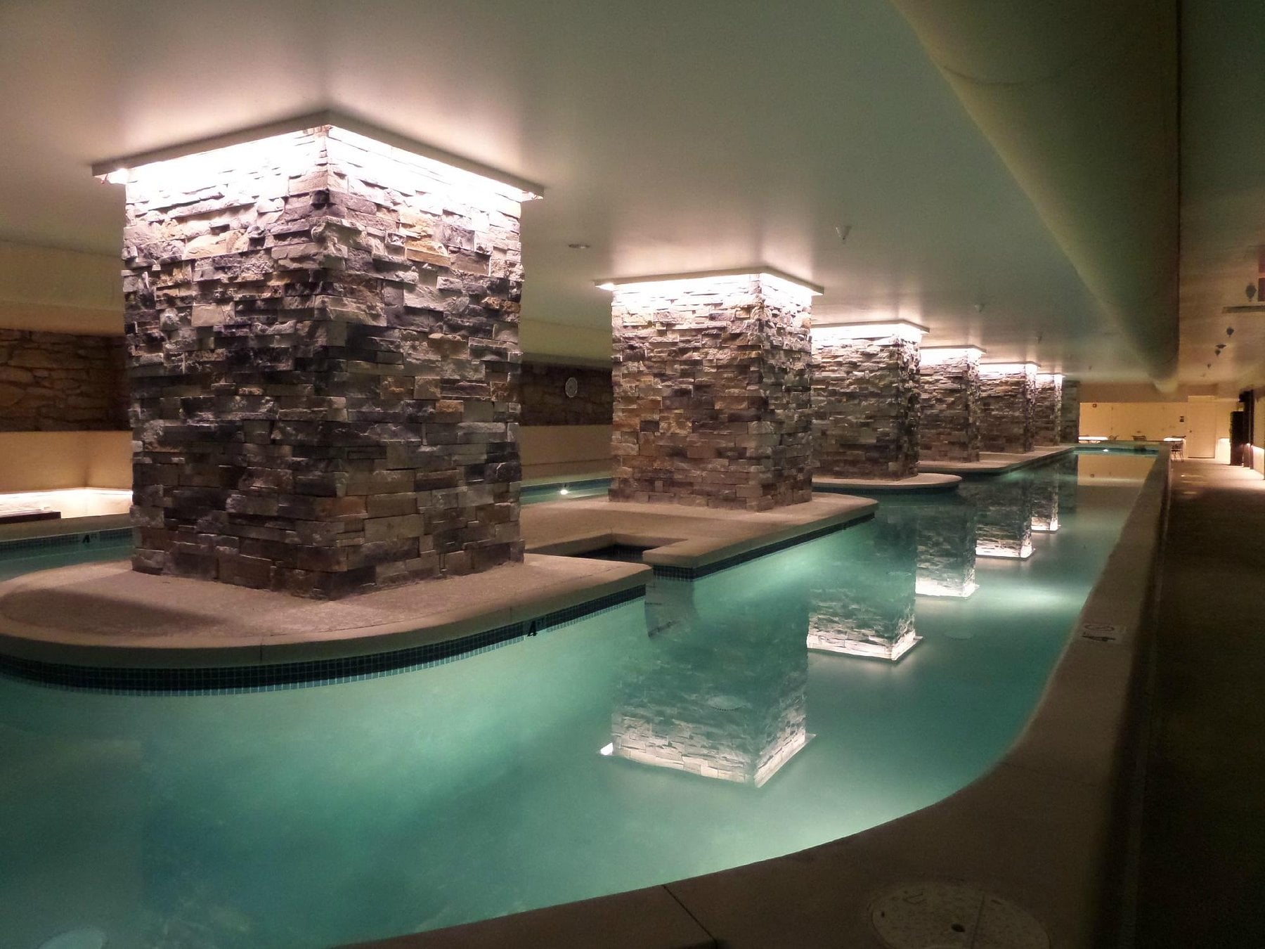 A modern indoor swimming pool at one of the finest Small-Town Luxury Hotels, with illuminated stone columns rising from the water and reflecting on the pool’s surface, creates a serene and elegant atmosphere.