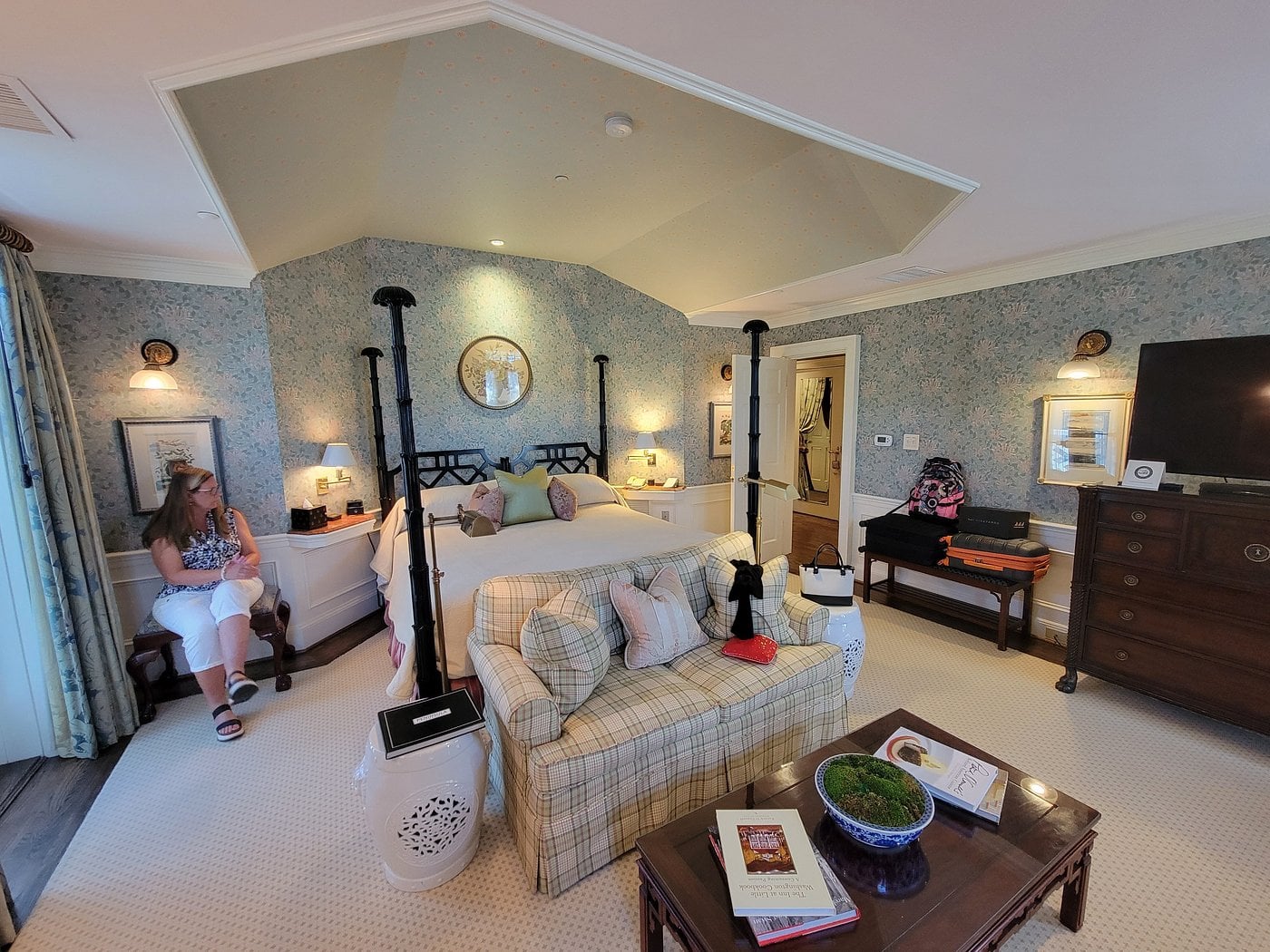 A cozy, well-lit bedroom in the style of small-town luxury hotels, with patterned wallpaper, a four-poster bed, a sofa at the foot of the bed, a woman by the window, and a table with books and a plant in the foreground.