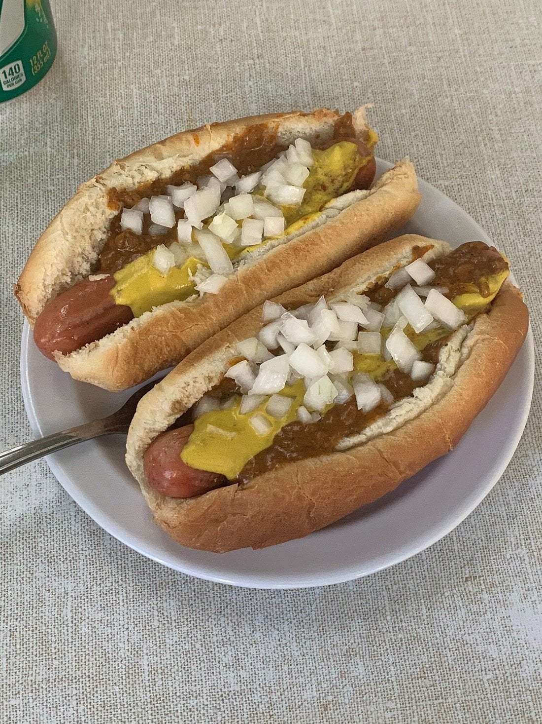 Two chili dogs topped with mustard and chopped onions sit on a white plate, perfect for classic road trip food stops. A fork rests nearby, alongside part of a green can, all set on a beige textured surface.