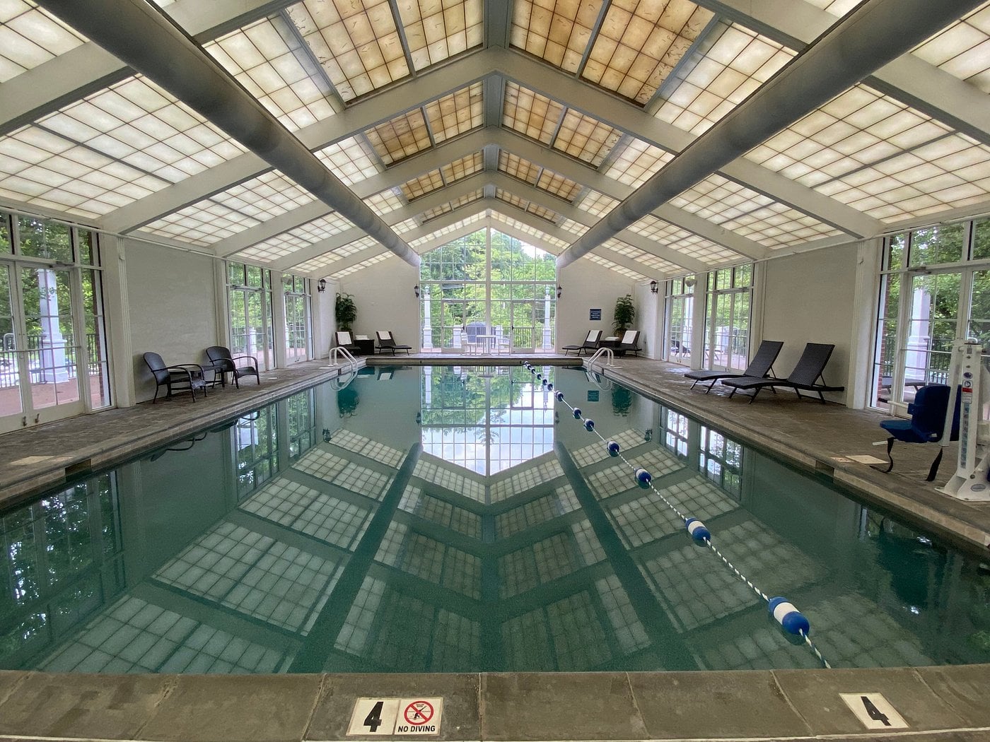 Indoor swimming pool with skylights and large windows, lounge chairs along both sides, and a lane divider in the water—a signature amenity found in Small-Town Luxury Hotels. Trees and outdoor furniture are visible through the windows at the far end.