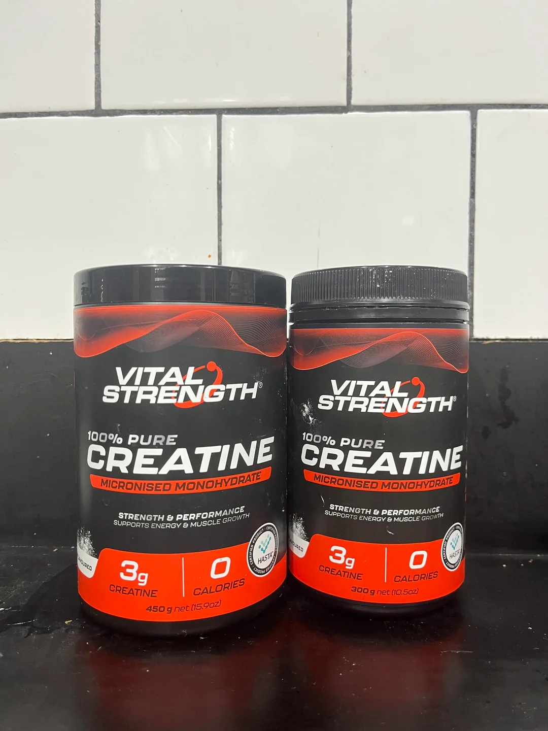 Two containers of Vital Strength 100% Pure Creatine, labeled “micronised monohydrate,” sit on a black countertop with a white tiled wall behind them. The containers have black lids and red and white labels.