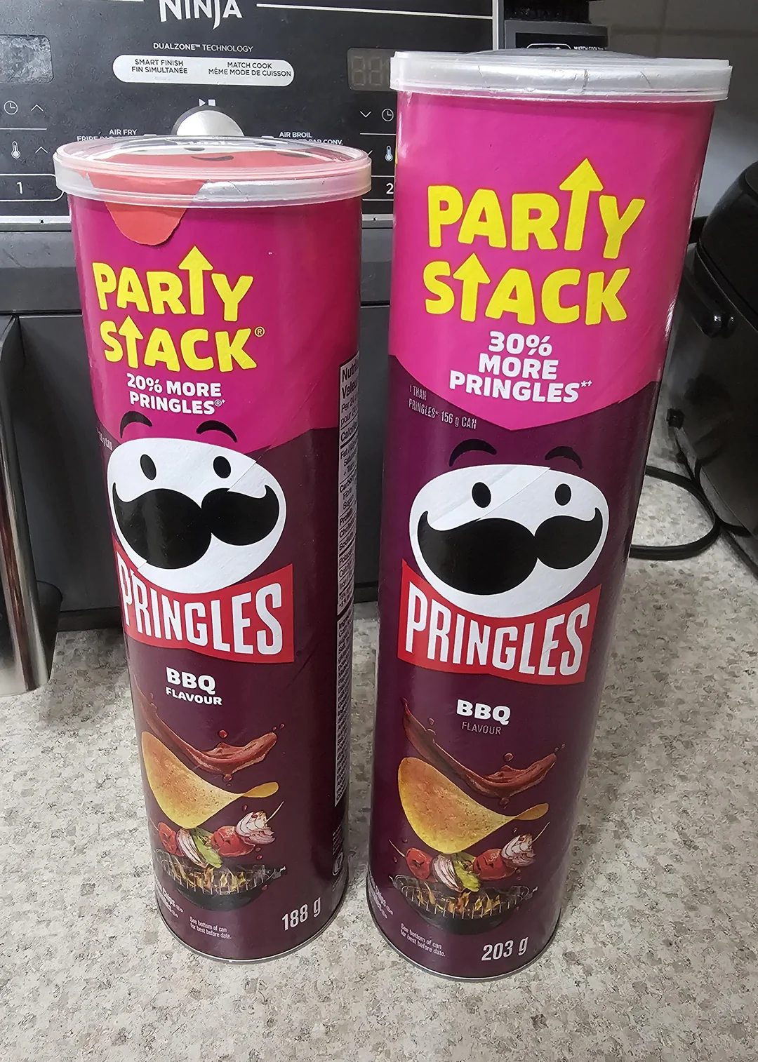 Two cans of Pringles BBQ flavour on a kitchen counter. One is labeled "PARTY STACK 20% MORE" (188g) and the other "PARTY STACK 30% MORE" (203g). A stove and blender are visible in the background.