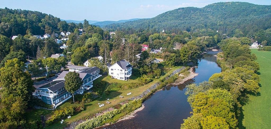 Aerial view of a unique American town nestled by the riverside, surrounded by lush green trees and hills, with houses and buildings near the water and open grassy fields.