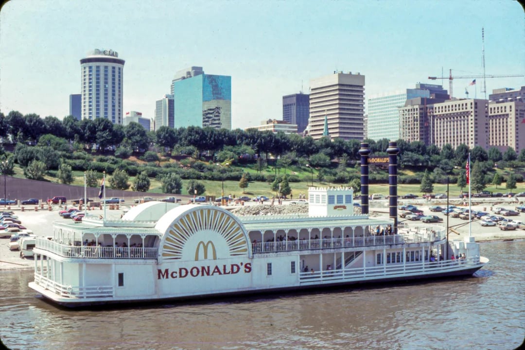 A McDonald's restaurant in the shape of a paddlewheel riverboat is docked on a river, with a city skyline, green park, and parked cars visible in the background—truly one of the Coolest McDonald's Locations.