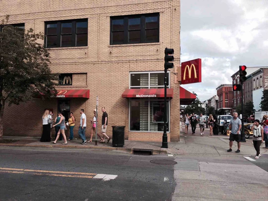People walk past a McDonald’s restaurant, one of the Coolest McDonald’s Locations, on a street corner with a brick building, red awnings, and two McDonald’s signs. The bustling street is filled with pedestrians and traffic lights.