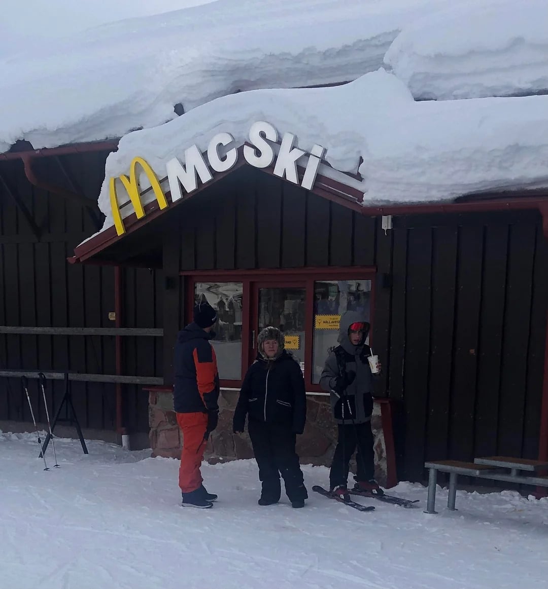 Three people in winter clothing stand outside "McSki," one of the Coolest McDonald's Locations, at a snowy ski resort. Snow piles on the roof while skis and snowboards rest nearby, and one person enjoys a drink cup in the frosty air.