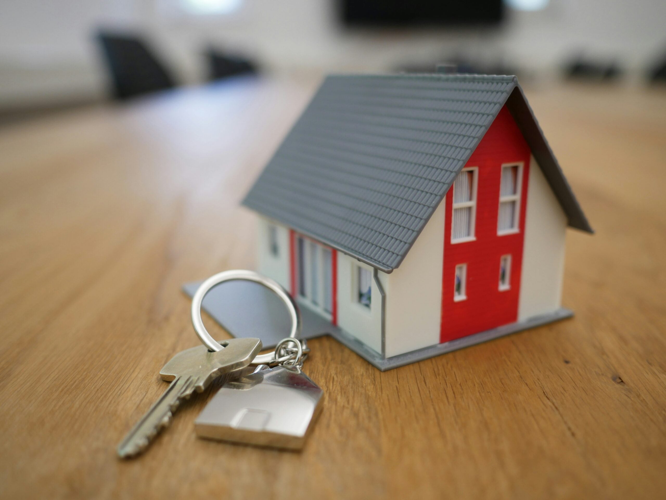 A set of house keys with a keychain lies on a wooden surface next to a small model of a house with a gray roof and red and white walls.