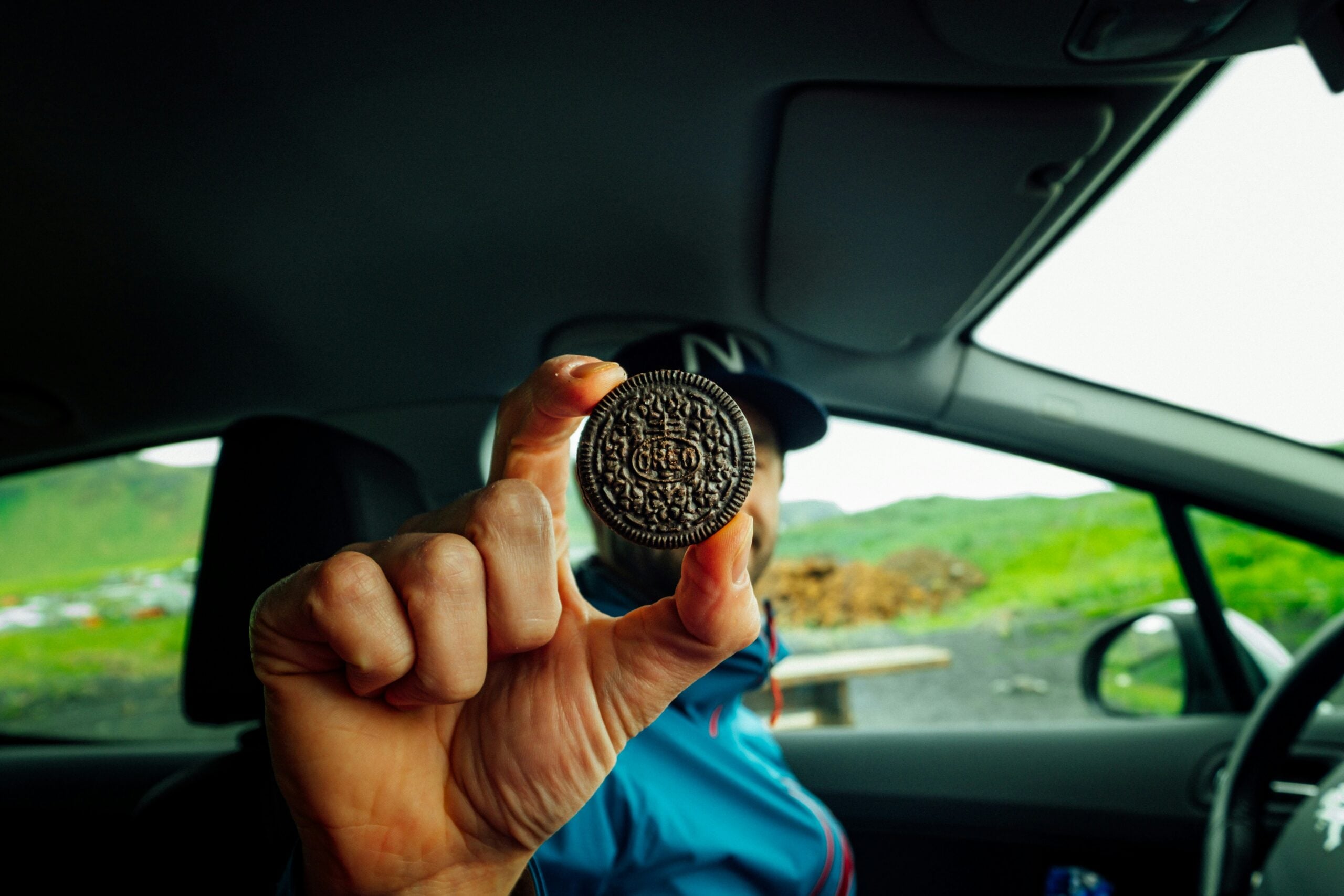 A person inside a car holds up a chocolate sandwich cookie close to the camera, obscuring their face. Green hills and a cloudy sky are visible through the car window in the background.