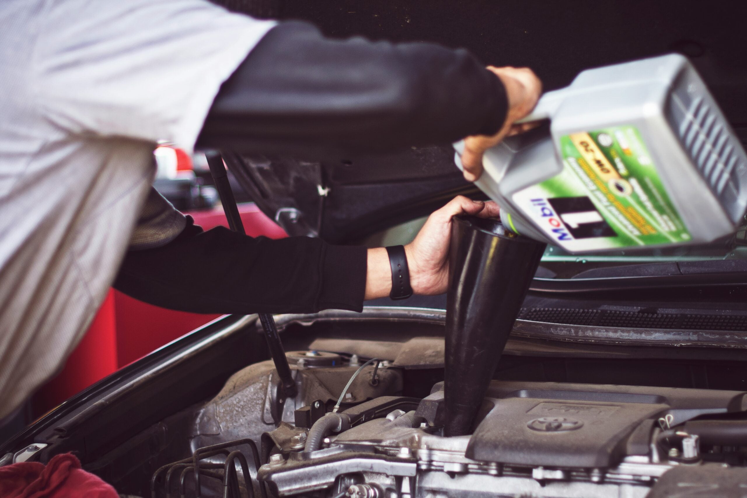 A person pours motor oil from a container into a car engine using a black funnel, with the car hood open and engine parts visible.