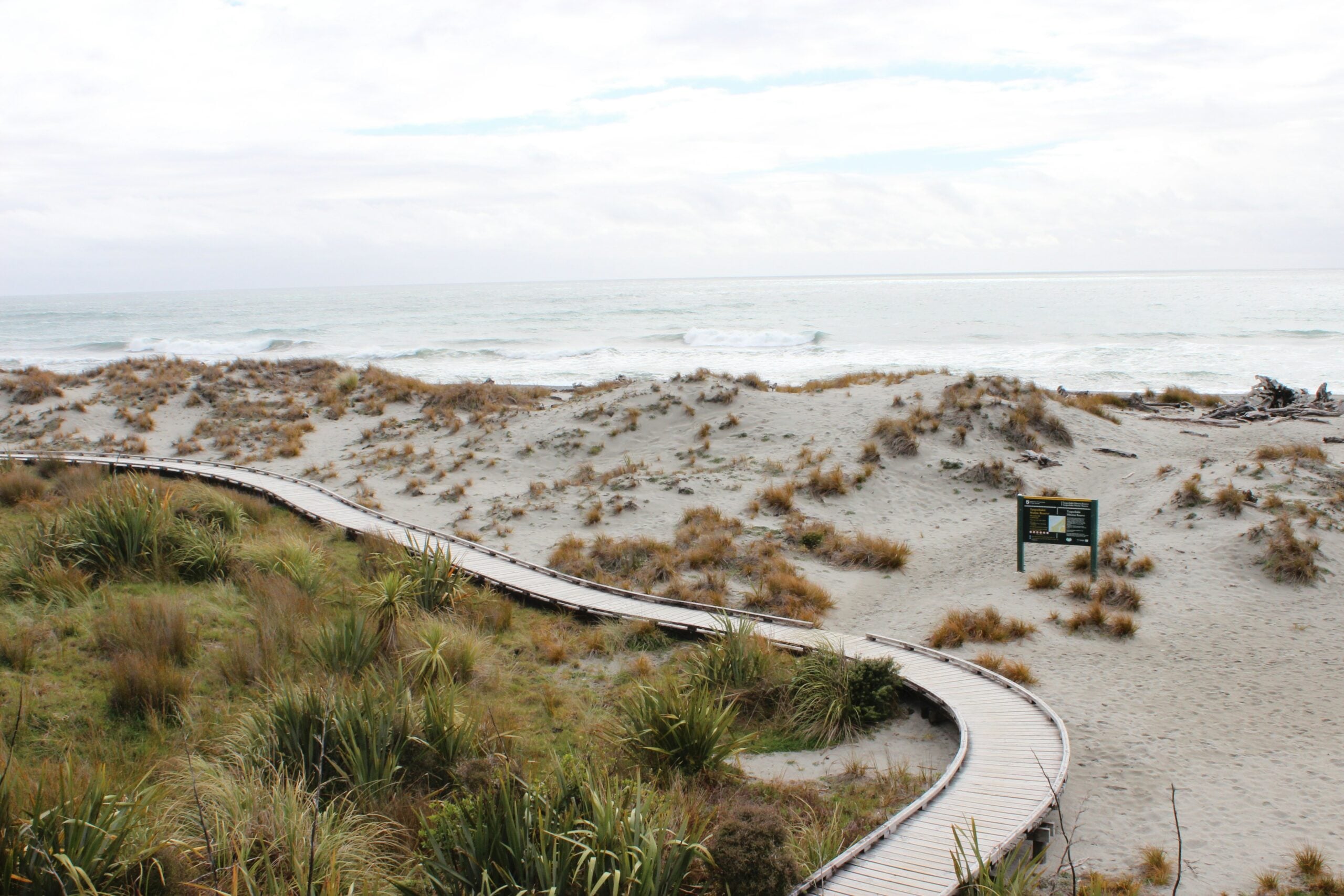 A winding wooden boardwalk leads through sandy dunes with scattered grasses toward the ocean under a cloudy sky. A small sign stands beside the path near the dunes.