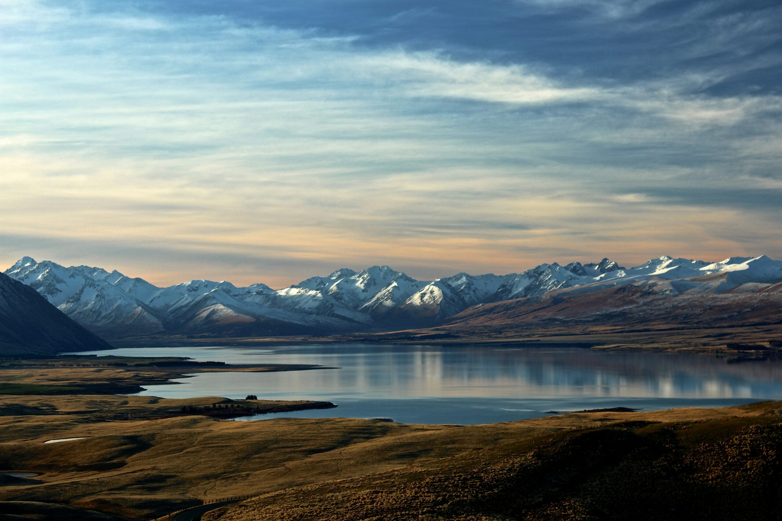 A serene lake stretches across the foreground, reflecting snow-capped mountains under a sky with soft, streaky clouds. Grassy plains surround the water, creating a peaceful, untouched landscape.