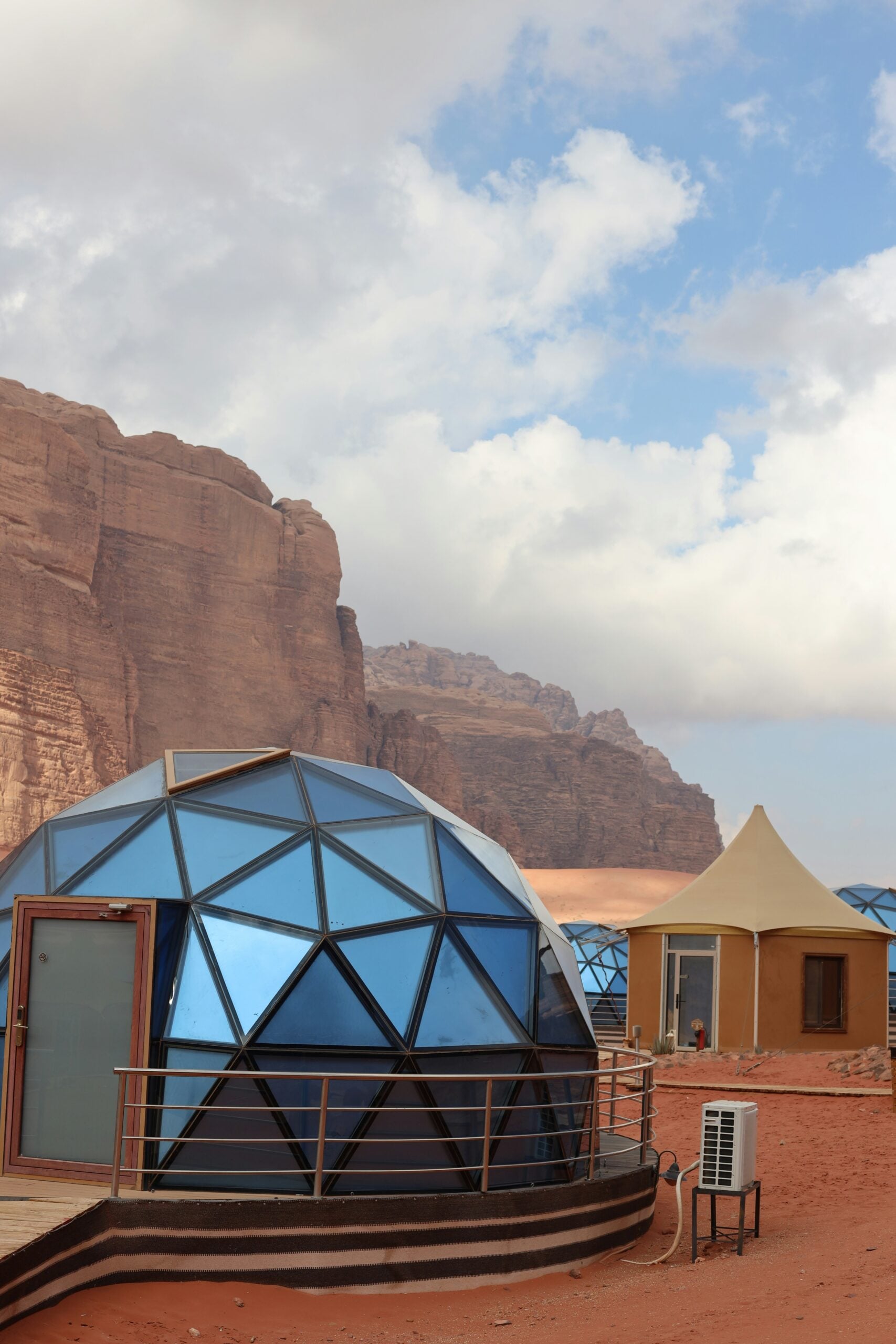 A geodesic dome with blue reflective panels and a beige tent stand on red desert sand, surrounded by rocky cliffs under a partly cloudy sky.