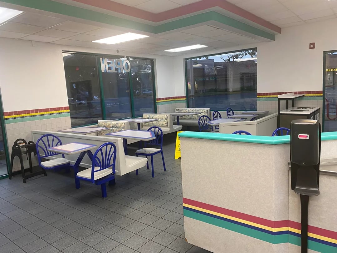 A brightly lit, empty fast-food restaurant interior with colorful trim, blue chairs, booth seating, tiled floors, and large windows. An "OPEN" sign is visible in the window and a caution sign is on the floor.
