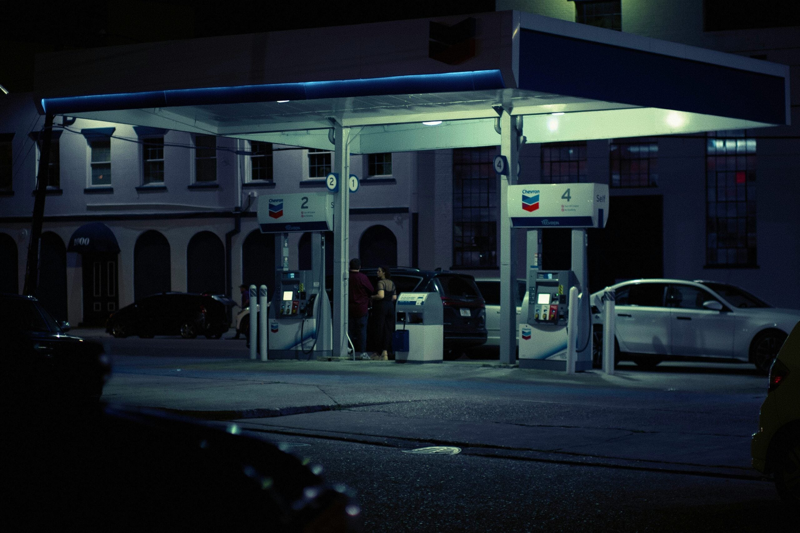A dimly lit Chevron gas station at night, with two cars parked at the pumps and people standing nearby. The scene has a moody, urban atmosphere, and surrounding buildings are visible in the background.