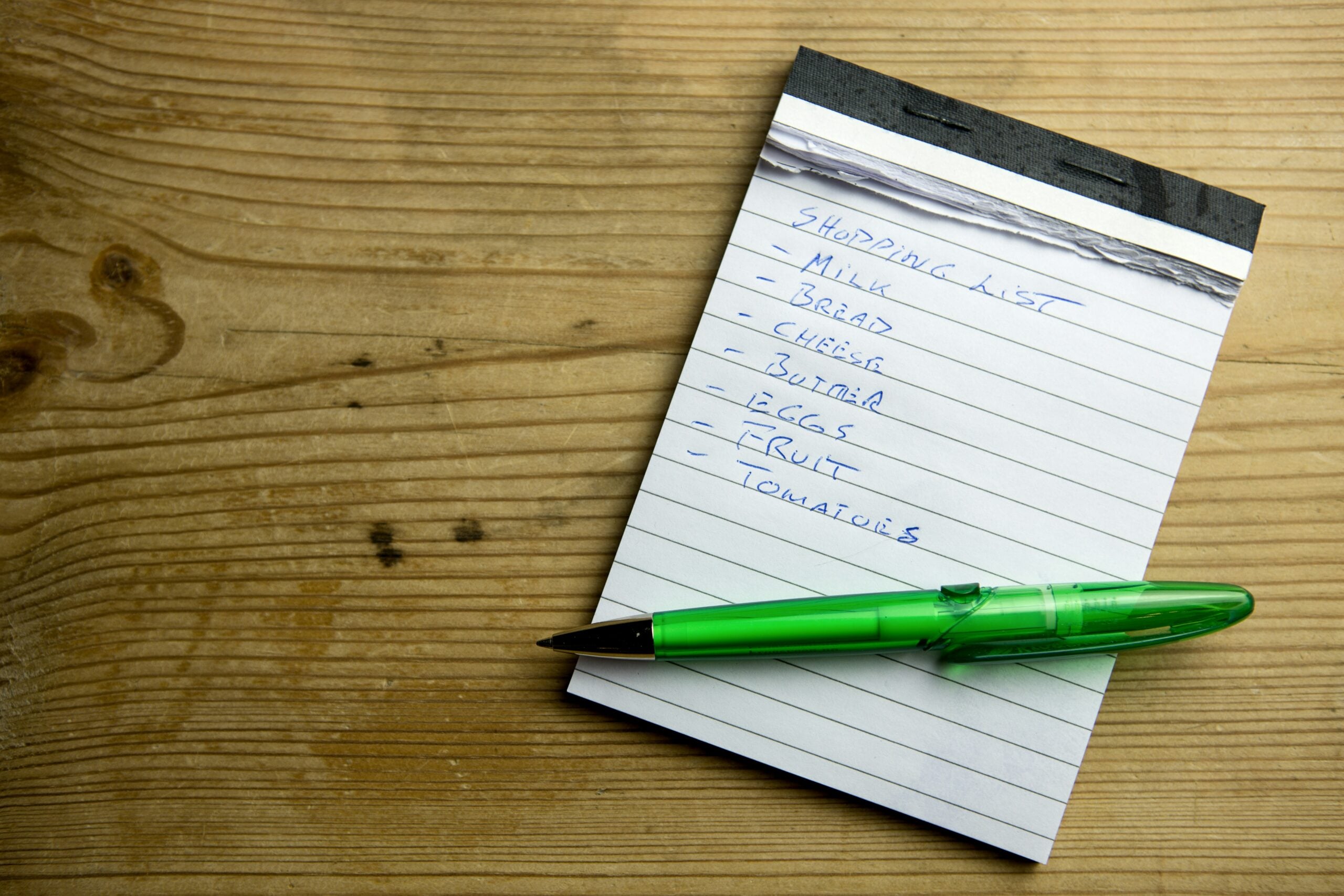 A green pen rests on a lined notepad with a handwritten shopping list that includes milk, bread, cheese, butter, eggs, fruit, and tomatoes, lying on a wooden surface.