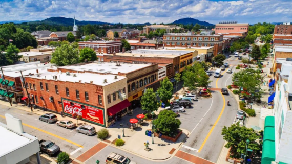 Aerial view of a lively small-town main street in one of the region&rsquo;s charming tourist towns, featuring historic brick buildings, shops, outdoor dining, parked cars, green trees, and distant hills under a partly cloudy sky.