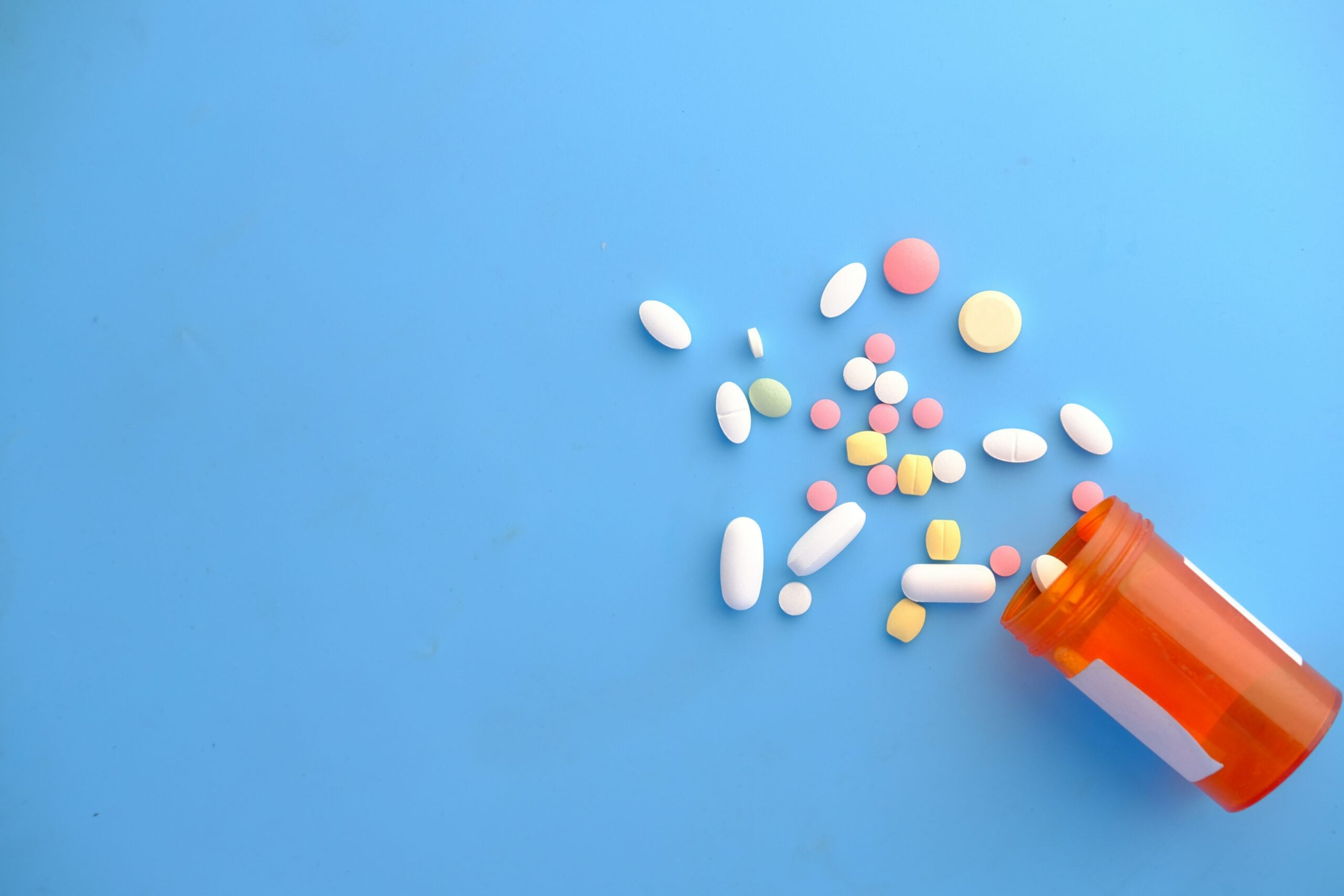 An orange prescription bottle tipped over on a blue surface, spilling out various colorful pills and tablets in different shapes and sizes.