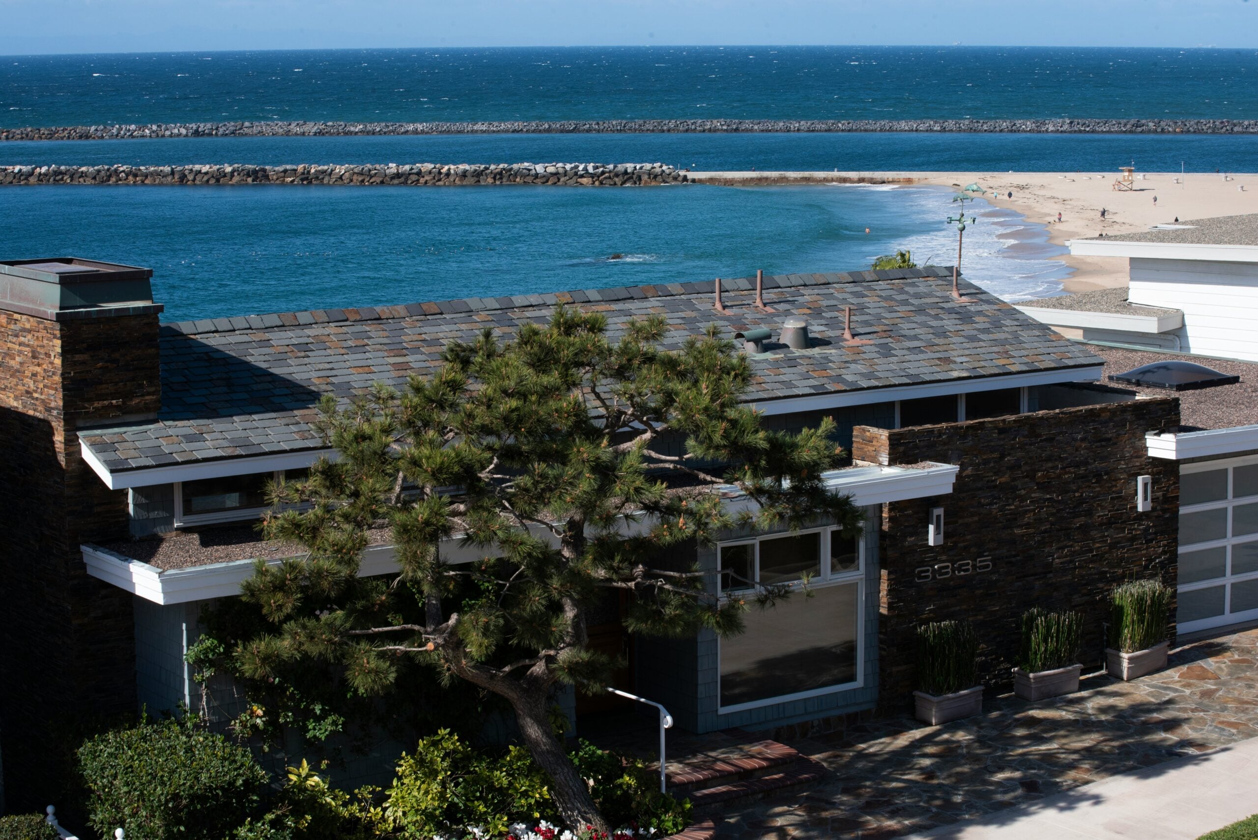 A modern house with a slate roof and stone accents sits by the ocean, with a sandy beach, blue water, breakwaters, and people in the distance under a clear sky.
