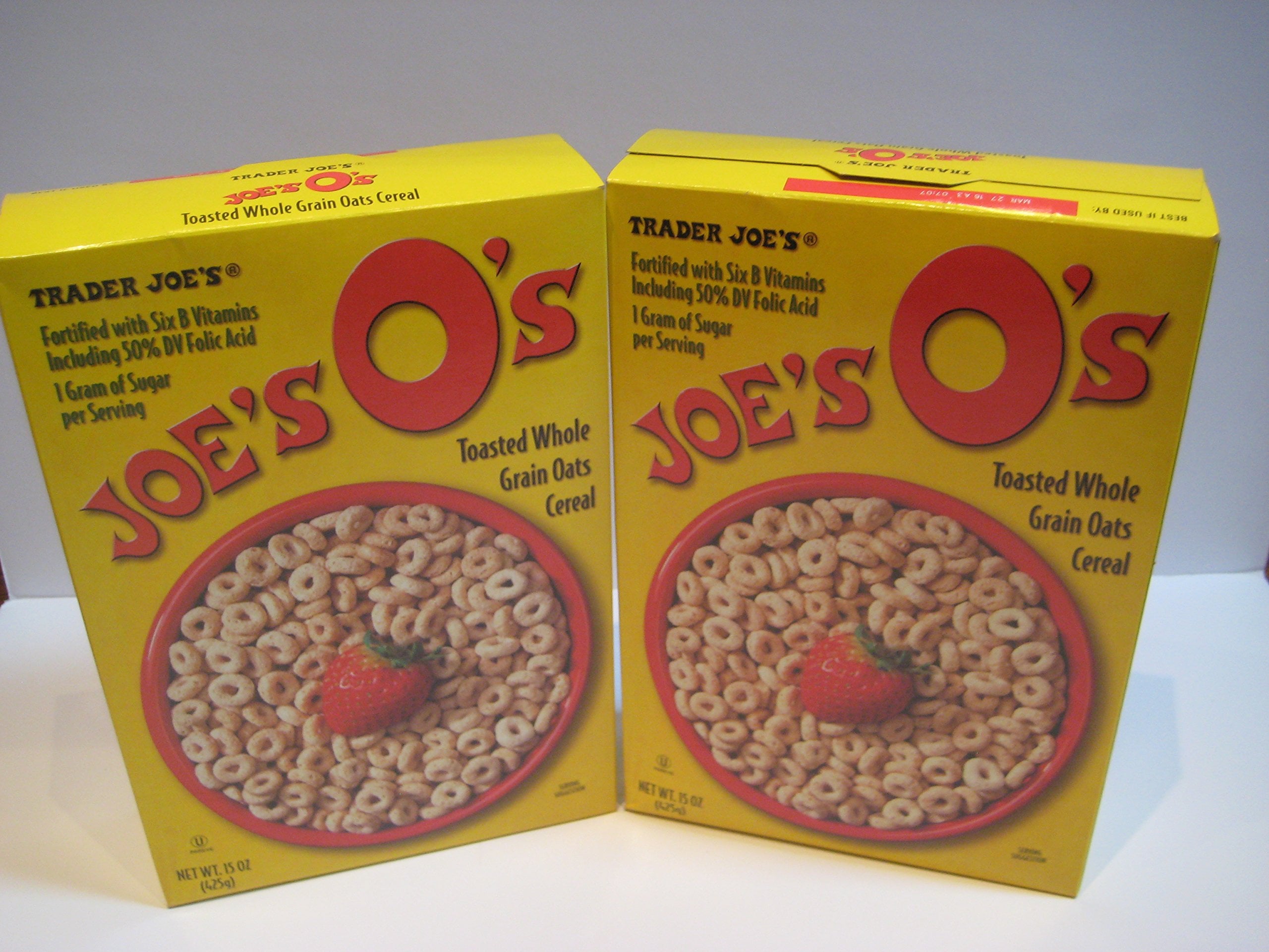 Two yellow boxes of Trader Joe’s Joe’s O’s Toasted Whole Grain Oats Cereal, each featuring a bowl of round oat cereal with a strawberry on the front, are standing side by side on a white surface.
