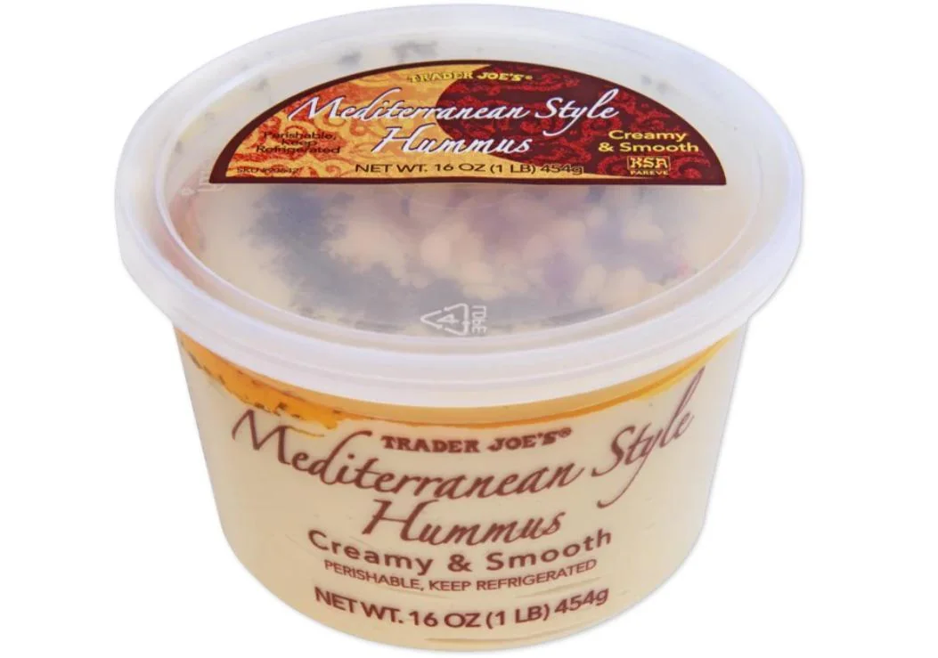 A 16-ounce container of Trader Joe's Mediterranean-Style Hummus, labeled "Creamy & Smooth," with a clear lid showing the tan hummus inside and colorful spices on top.