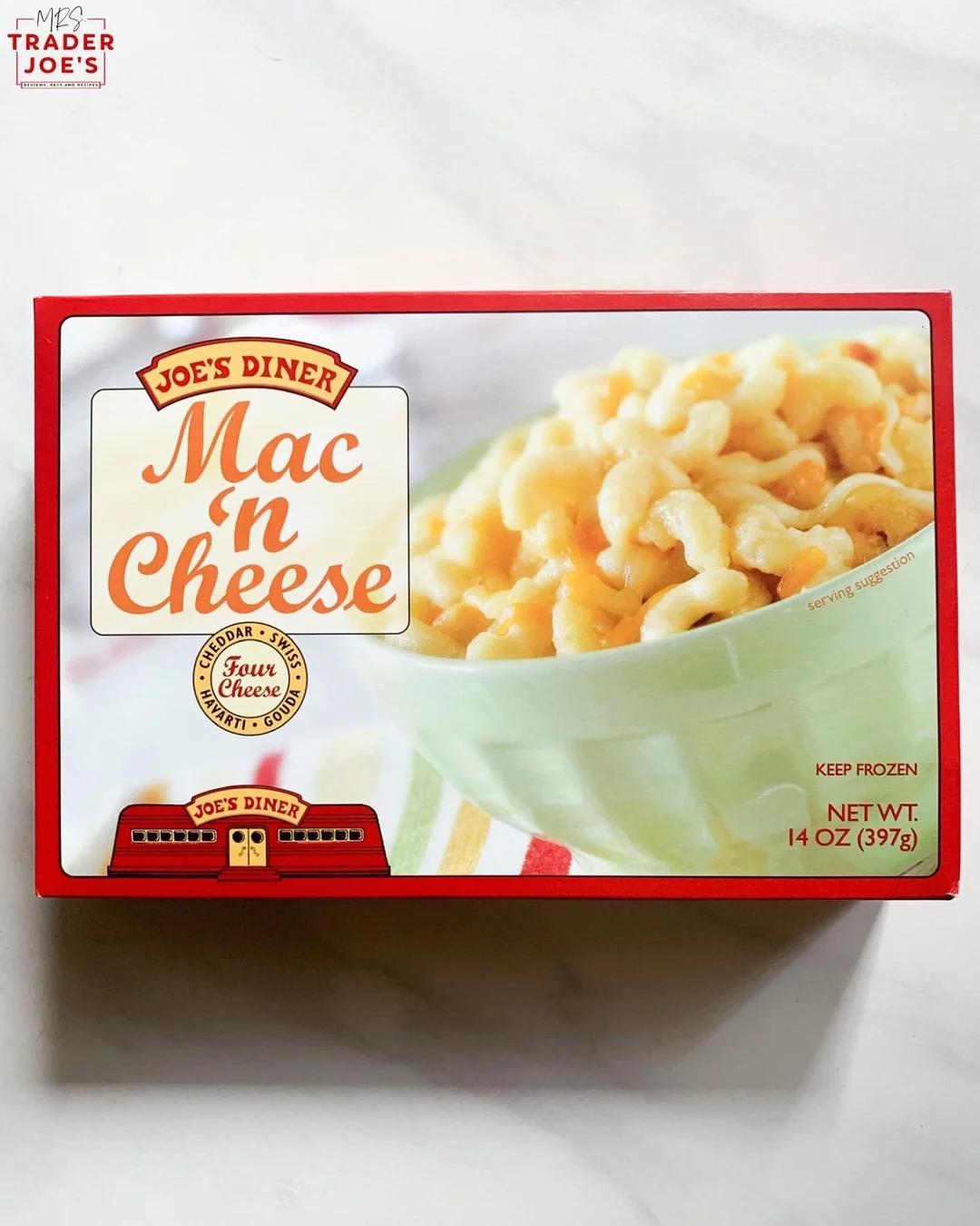 A box of Trader Joe’s Joe’s Diner Mac ‘n Cheese is shown, featuring a bowl of macaroni and cheese on the front. The packaging highlights "Faux Cheddar" and notes the net weight as 14 oz (397g).