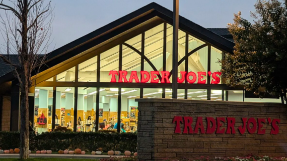 A Trader Joe&rsquo;s store with a peaked roof and large windows is shown at dusk. The bright red sign welcomes shoppers, showcasing the charm often found at Trader Joe's new store openings, with pumpkins and bushes lining the walkway in front.