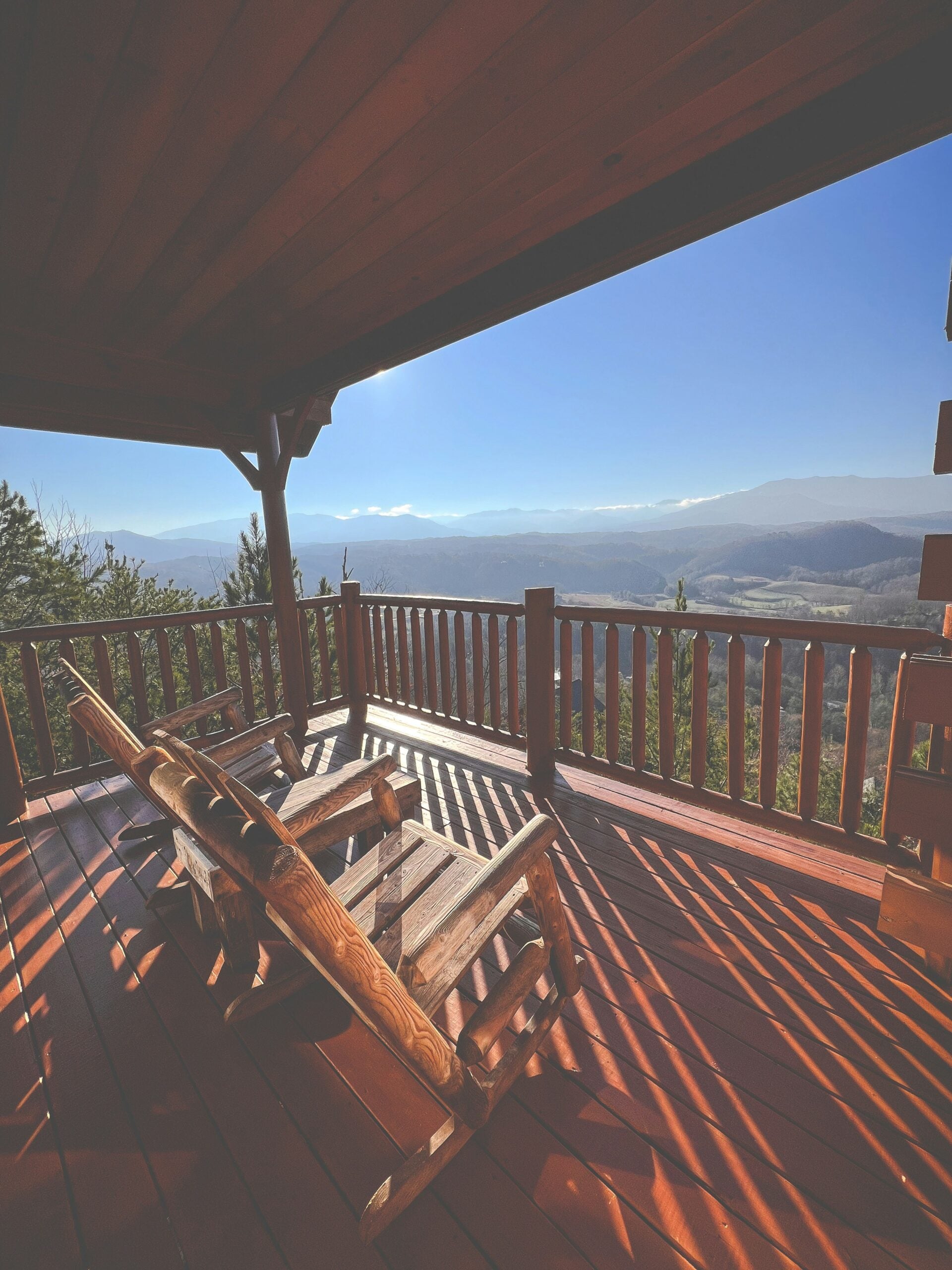 A wooden porch with two rustic rocking chairs overlooks a scenic mountain landscape under a clear blue sky, with sunlight casting shadows across the deck.
