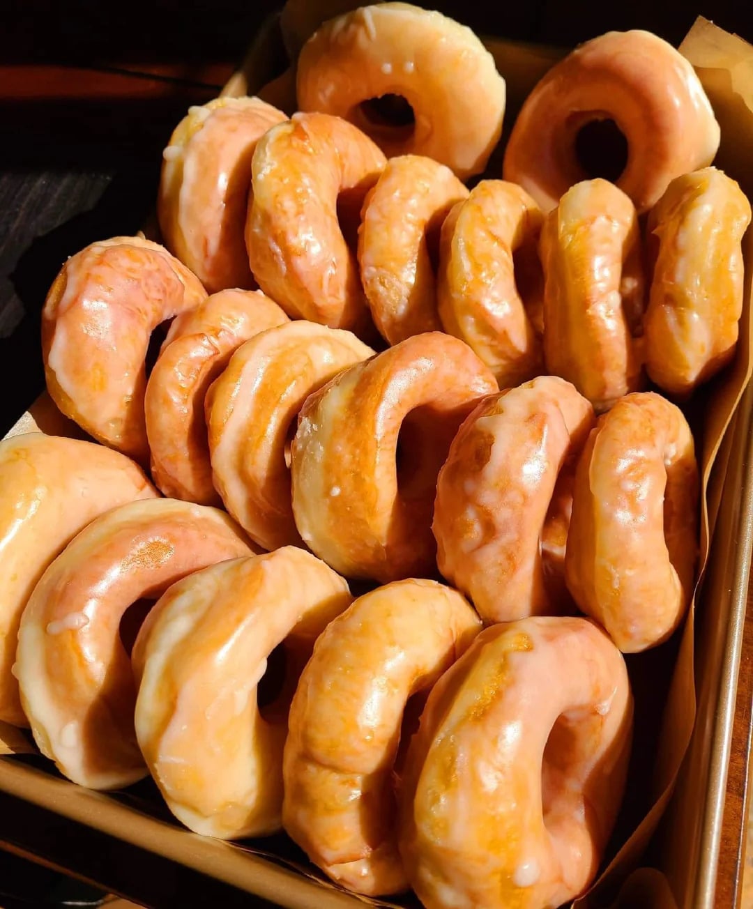 A box filled with several shiny, golden-brown glazed donuts, expertly arranged in rows under bright lighting—these irresistible fast-food desserts are perfect for any sweet craving.