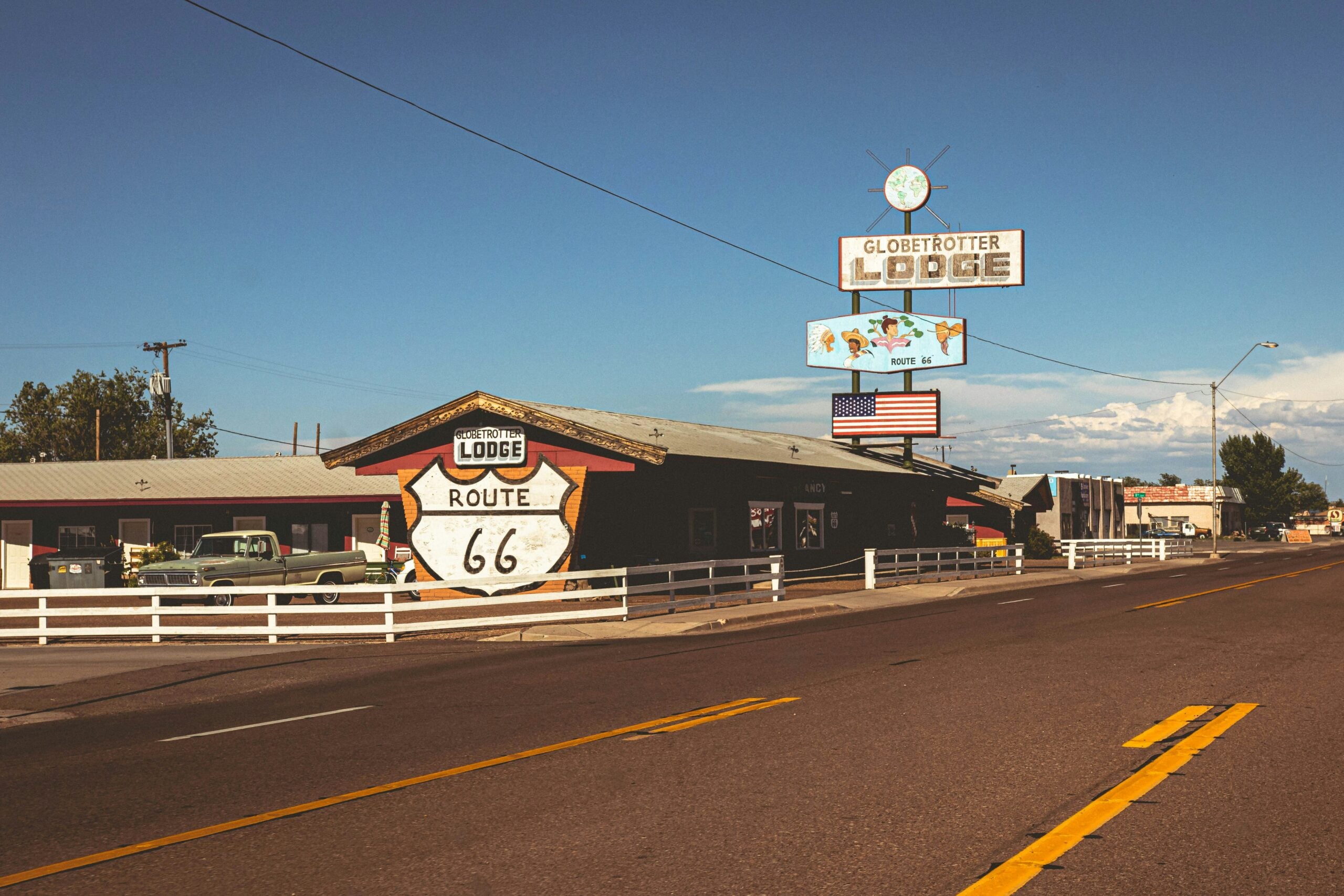 A retro-style lodge with a Route 66 emblem on its exterior sits along a sunny roadside, featuring a vintage neon sign and an American flag; classic cars and a blue sky complete the scene.