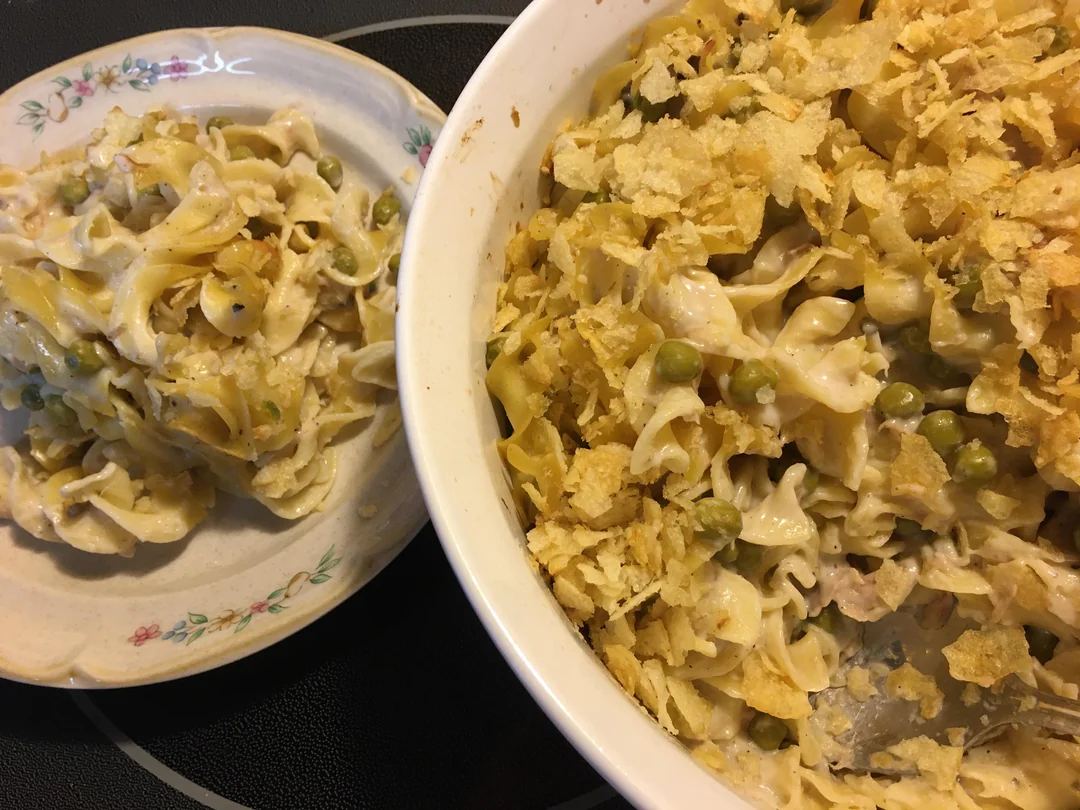 A serving of tuna noodle casserole with peas and crushed potato chips on top is on a floral plate next to a large baking dish filled with the same casserole.
